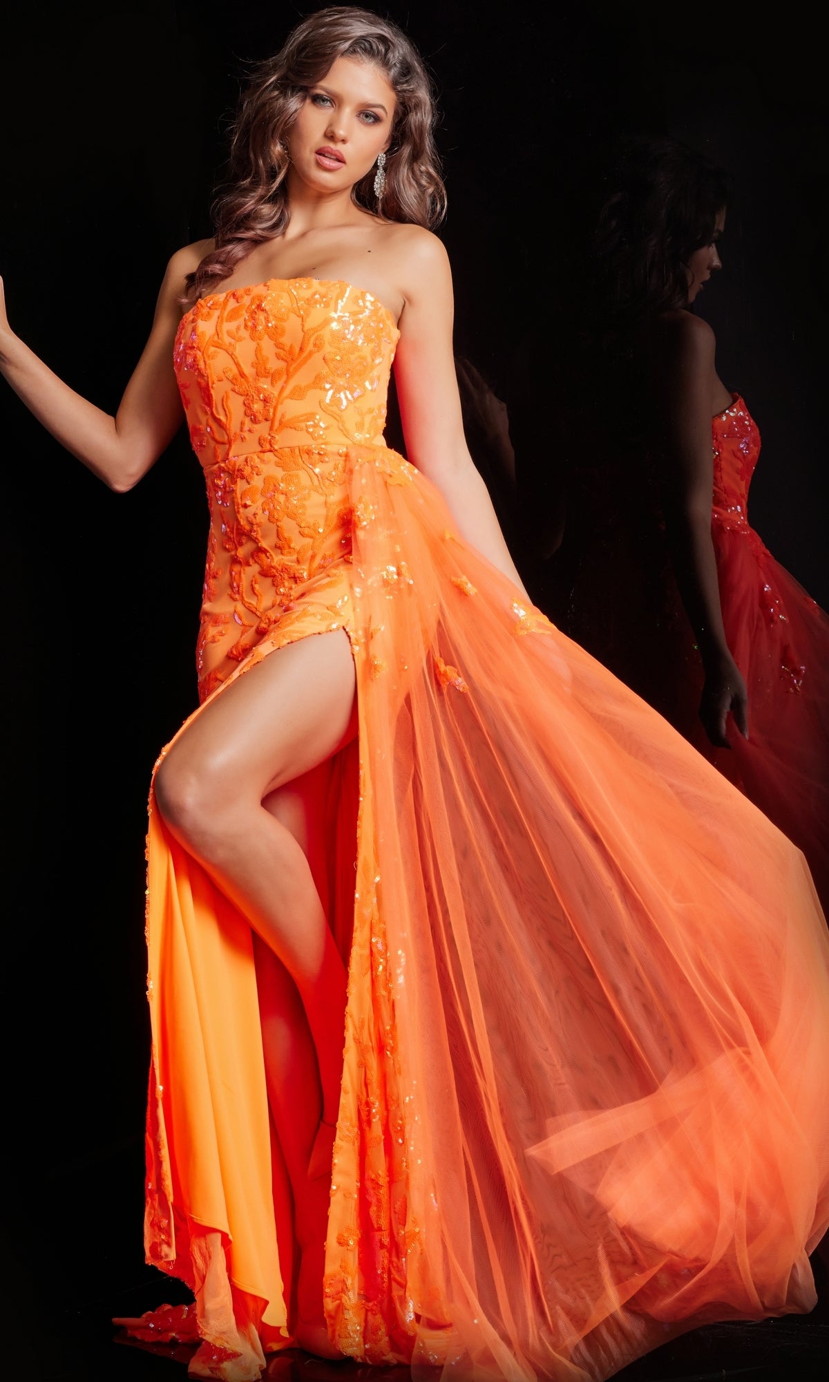A woman poses in the Jovani Strapless Long Neon Sequin Prom Dress 26134, featuring a sheer tulle overlay and high slit. Her reflection glows against a dark background as the dress dazzles in bright orange sequins and shimmering accents.